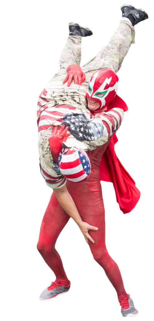 A wrestler in a red mask, unitard and cape body slamming another wrestler wearing an American flag mask, shirt and camouflage pants.