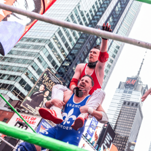 A man in blue singlet holds another man in a red singlet and white pants on his shoulders in a wrestling ring with city buildings in the background.