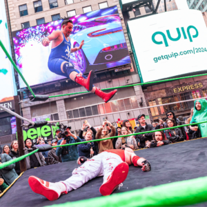 A man in blue singlet jumps from top rope of a wrestling ring with his legs extended in front of him, with another man on the ground.