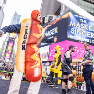 A woman dressed in a hot dog outfit with the word "Vegan" written on it in vertical, black lettering, stands in a wrestling ring.