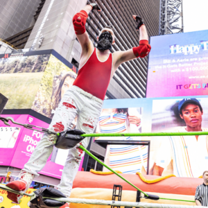 A man in a red singlet top and white plants stands on the middle ropes in a wrestling ring gesturing to the crowd.