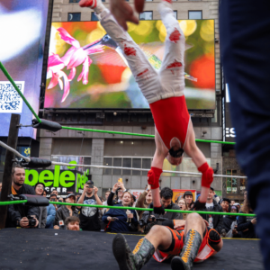 A wrestler with a mohawk wearing white pants and a red singlet top flips backwards onto another wrestler laying on the ground of a wrestling ring.