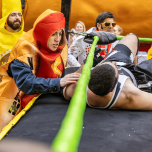 A woman dressed in a hot dog outfit with the word "Vegan" written on it in vertical, black lettering, stands in a wrestling ring.