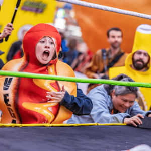 A woman dressed in a hot dog outfit with the word "Vegan" written on it in vertical, black lettering, stands in a wrestling ring.