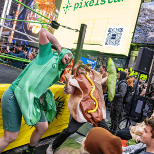 A man dressed like a pickle and another man dressed like a hot dog pose comically outside of a wrestling ring for a crowd.