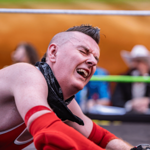 A male wrestler sporting a mohawk in red gear looks in agony, while laying on his belly in a wrestling ring.