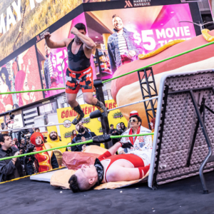 A male wrestler jumps off the corner of a wrestling ring onot another male wrestler laid out on the ground.