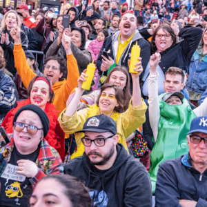 Among a crowd of people, three women are dressed in colors representing different condiments.